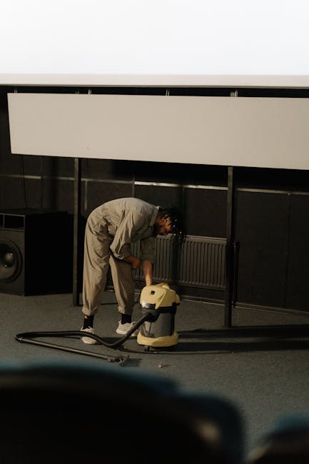 A professional cleaner wearing a light gray uniform is bent over on a dark carpeted floor using a yellow vacuum or carpet cleaning machine, which is connected to a long black hose. The background feat