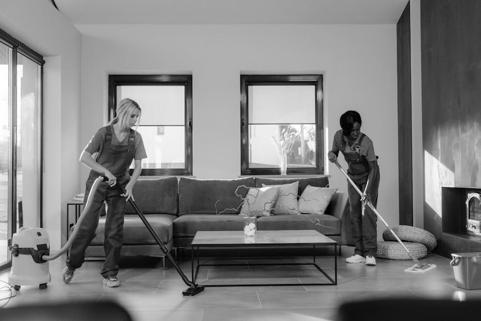 A professional cleaner wearing a light gray uniform is bent over on a dark carpeted floor using a yellow vacuum or carpet cleaning machine, which is connected to a long black hose. The background feat