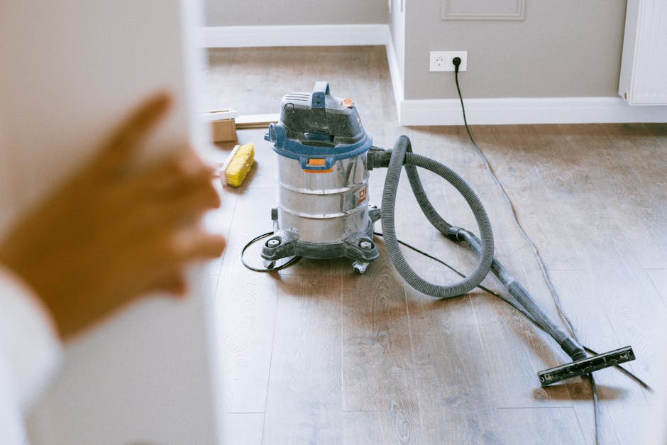 A commercial cleaning vacuum cleaner with a metal body, black and orange accents, and a long black hose attached to a cleaning nozzle is positioned on a wooden floor in a residential room. The room fe